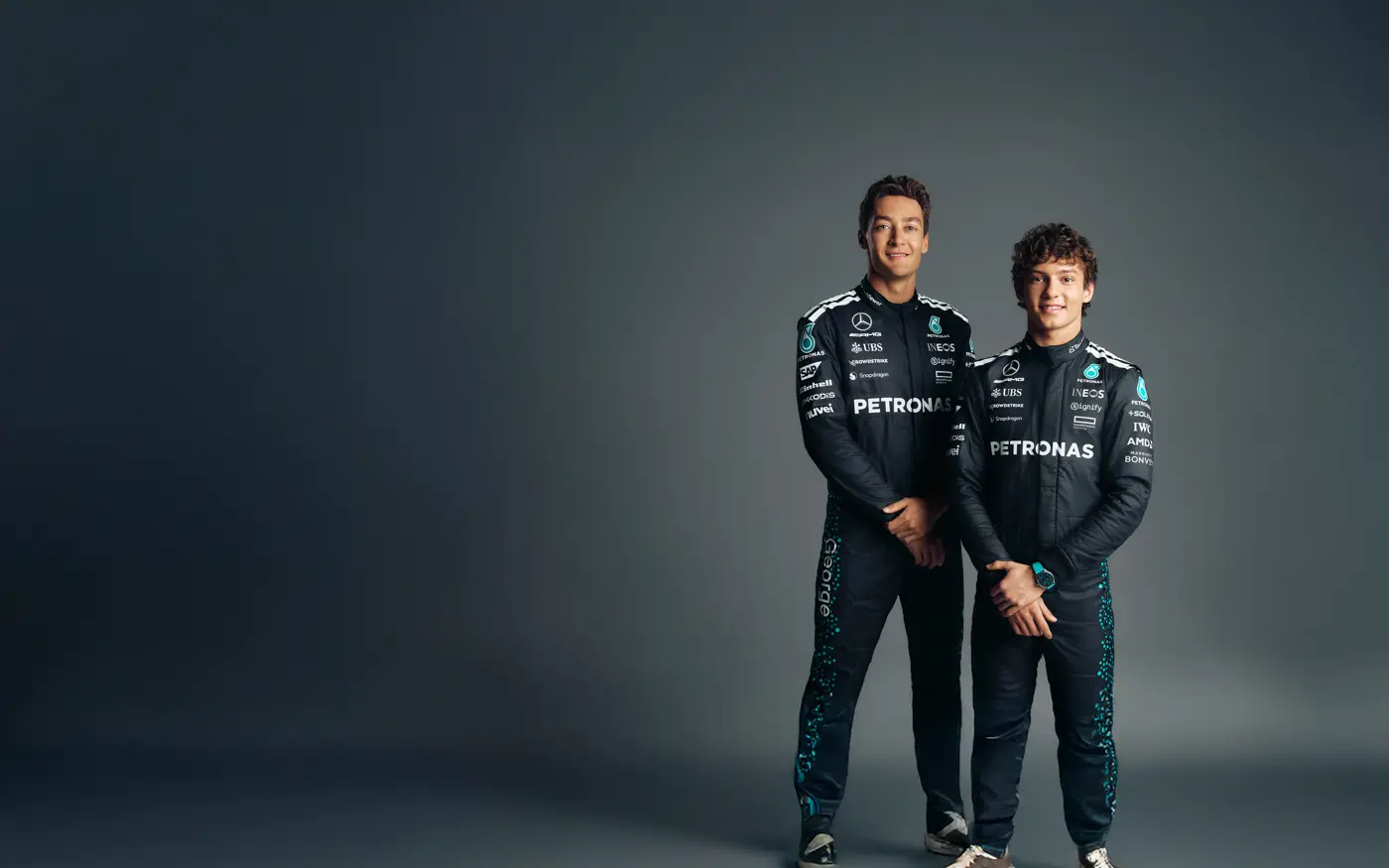 Two Mercedes-AMG Petronas Formula One drivers in full race suits posing against a dark studio background, smiling confidently with arms relaxed.