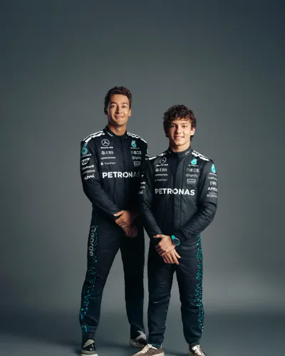 Two Mercedes-AMG Petronas Formula One drivers in full race suits posing against a dark studio background, smiling confidently with arms relaxed.