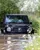 Black Mercedes-Benz G-Class SUV driving through deep muddy water during an off-road experience, surrounded by dense greenery and wildflowers.