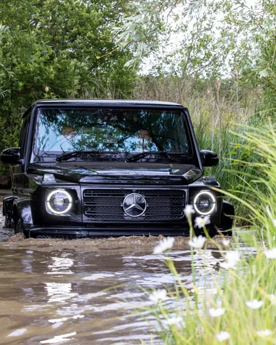 Black Mercedes-Benz G-Class SUV driving through deep muddy water during an off-road experience, surrounded by dense greenery and wildflowers.