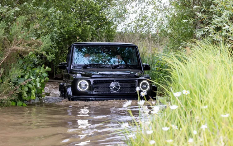 Black Mercedes-Benz G-Class SUV driving through deep muddy water during an off-road experience, surrounded by dense greenery and wildflowers.