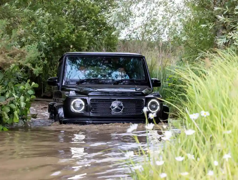 Black Mercedes-Benz G-Class SUV driving through deep muddy water during an off-road experience, surrounded by dense greenery and wildflowers.