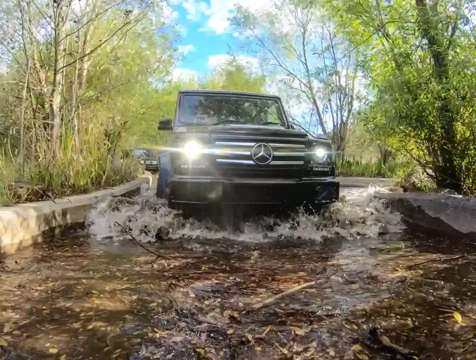 A modern black car parked on a wet driveway in front of a house, with reflections on the ground and greenery surrounding the scene.