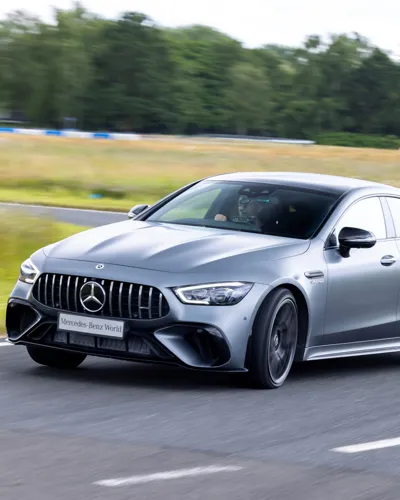 A silver Mercedes-AMG speeding on a test track at Mercedes-Benz World, with the driver focused and blurred trees and grass in the background.