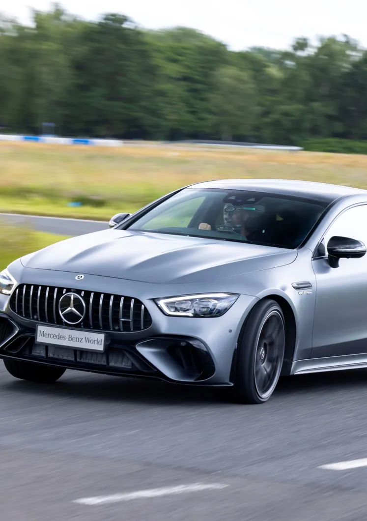 A silver Mercedes-AMG speeding on a test track at Mercedes-Benz World, with the driver focused and blurred trees and grass in the background.
