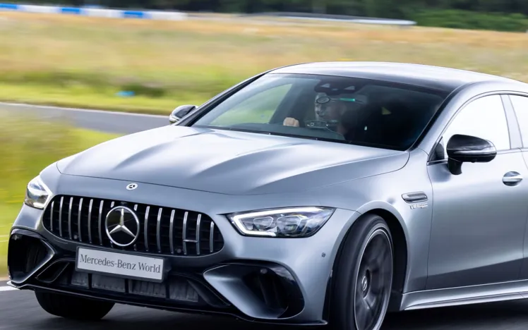 A silver Mercedes-AMG speeding on a test track at Mercedes-Benz World, with the driver focused and blurred trees and grass in the background.