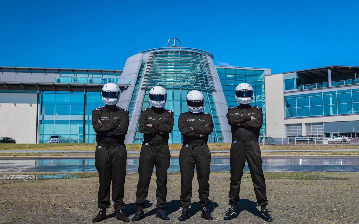 Four professional drivers in black racing suits and helmets standing with arms crossed in front of the Mercedes-Benz World building under a clear blue sky.