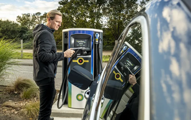 A man prepares to plug a charging cable into a black Mercedes-Benz electric vehicle at an outdoor charging station, with greenery and trees in the background.