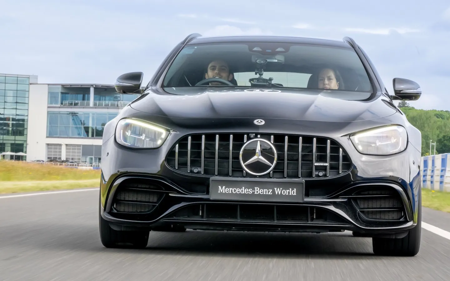 A black Mercedes-Benz AMG drives on a track at Mercedes-Benz World, with two people visible inside the vehicle and a modern glass building in the background.