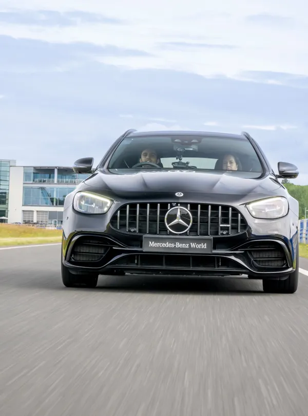 A black Mercedes-Benz AMG drives on a track at Mercedes-Benz World, with two people visible inside the vehicle and a modern glass building in the background.