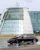 A black Mercedes-Benz EQE electric car drives through water on a test track in front of the iconic glass-fronted Mercedes-Benz World building.