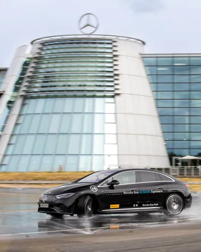 A black Mercedes-Benz EQE electric car drives through water on a test track in front of the iconic glass-fronted Mercedes-Benz World building.