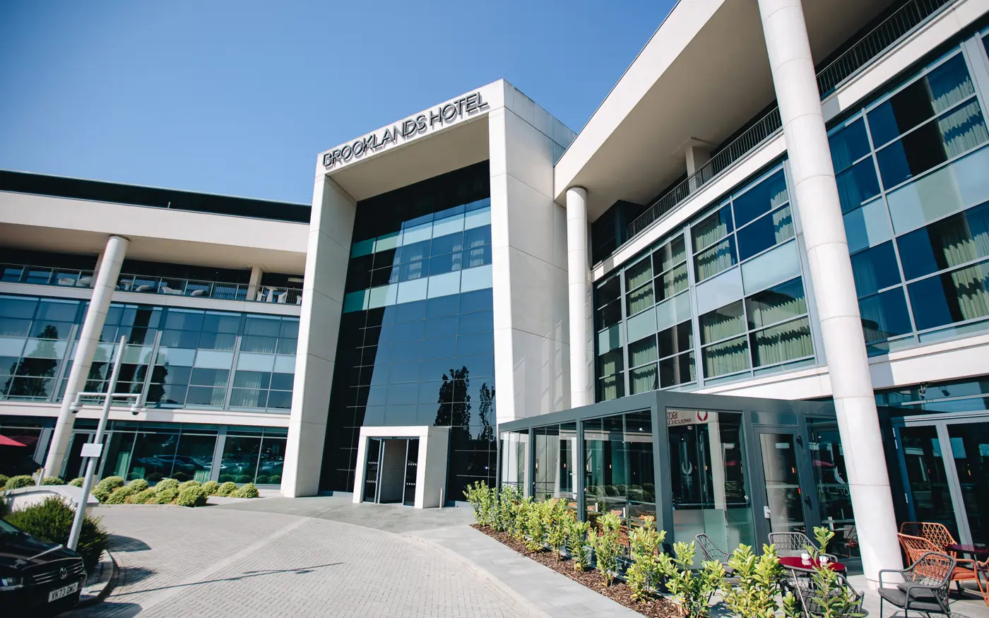 Exterior view of Brooklands Hotel, showcasing its modern glass-fronted architecture, tall white columns, and landscaped entrance under a clear blue sky.