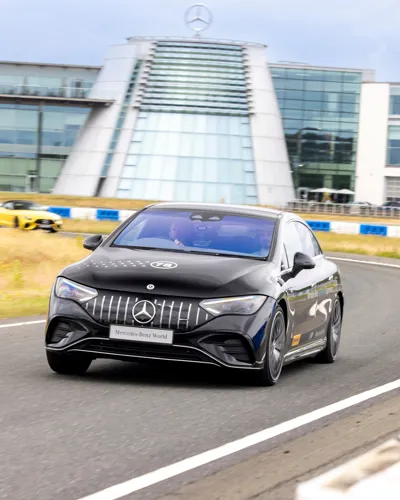 A black Mercedes-Benz car speeds along a track outside Mercedes-Benz World, with the building's iconic glass façade and logo visible in the background.
