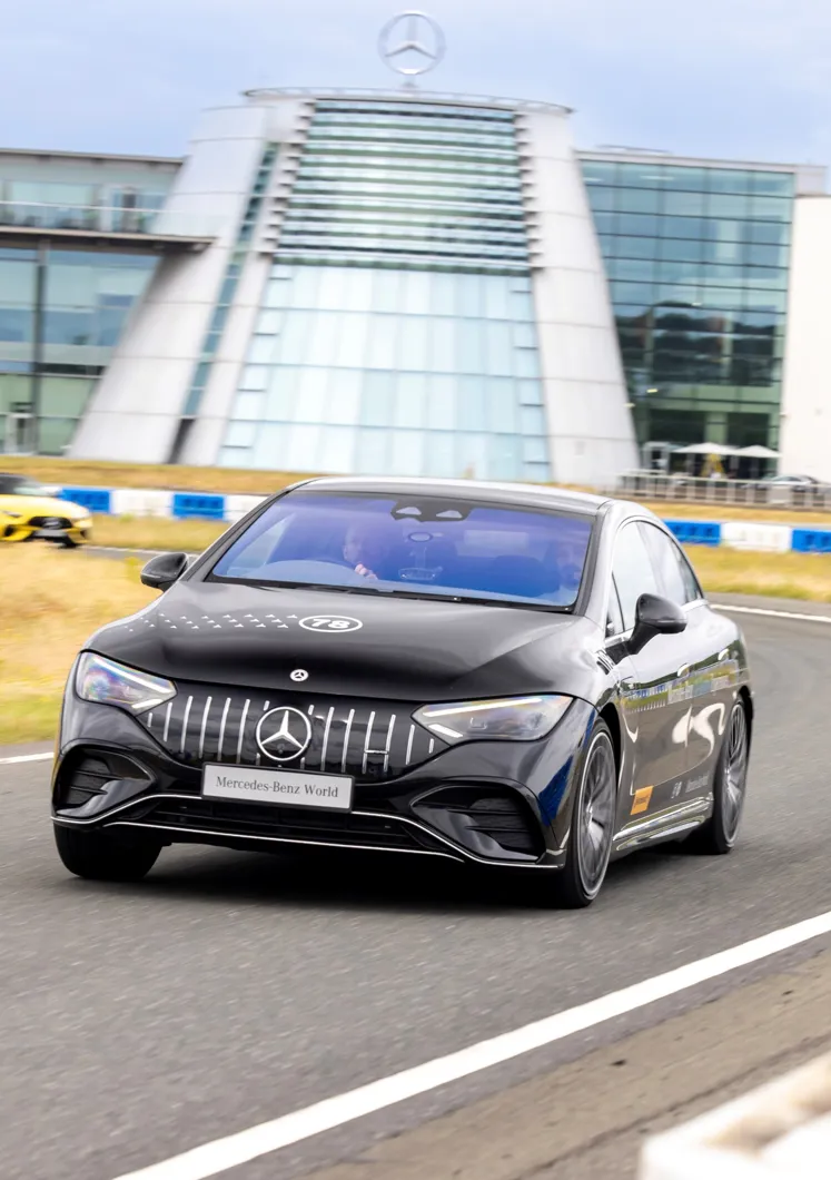 A black Mercedes-Benz car speeds along a track outside Mercedes-Benz World, with the building's iconic glass façade and logo visible in the background.