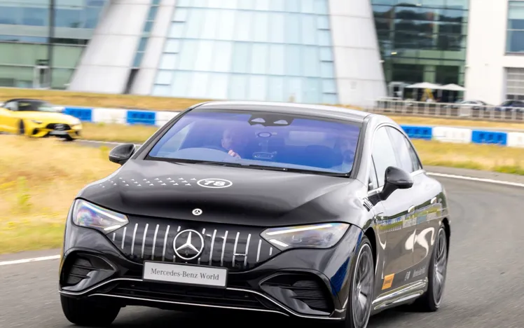 A black Mercedes-Benz car speeds along a track outside Mercedes-Benz World, with the building's iconic glass façade and logo visible in the background.