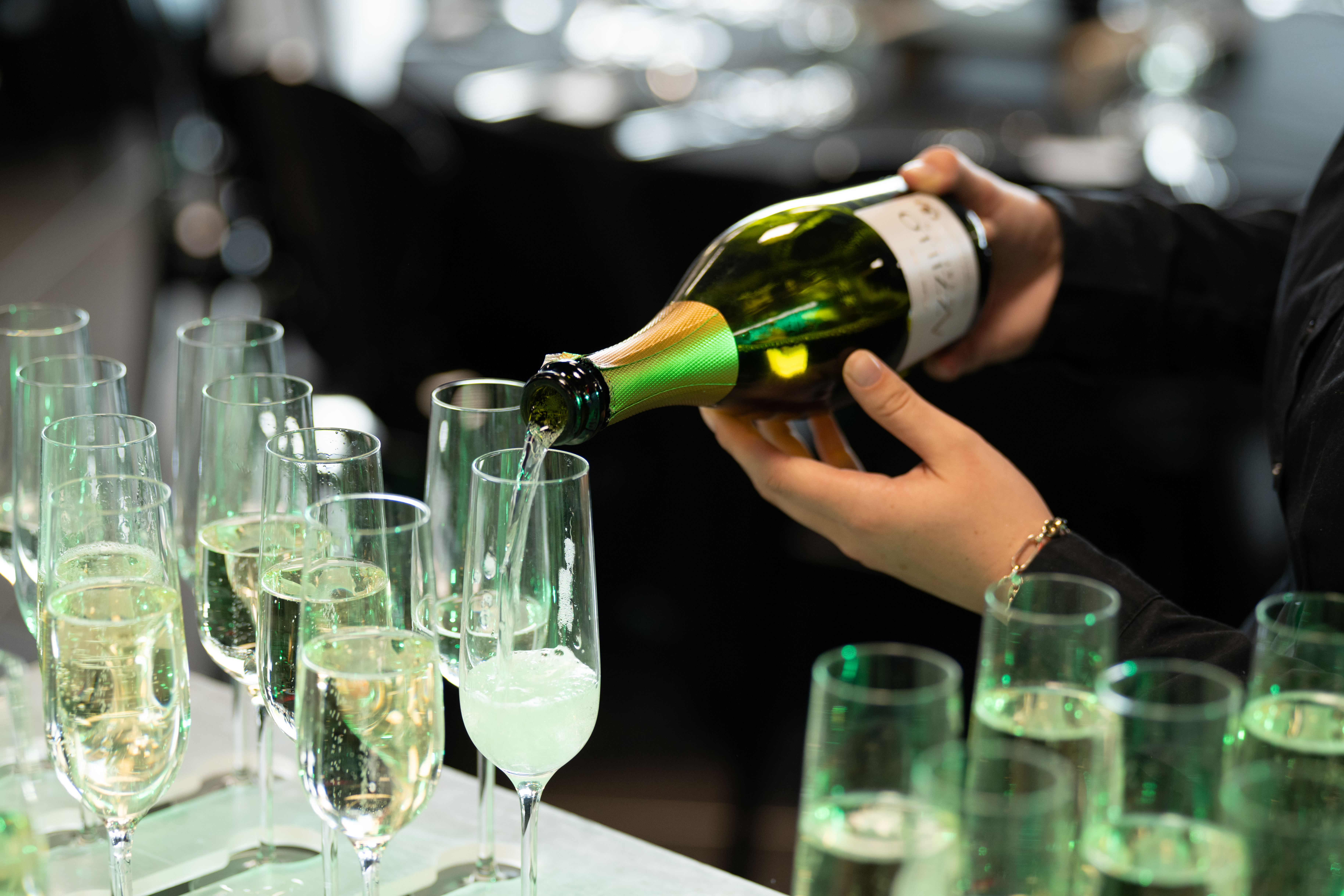 A hand pours white wine or champagne into a row of tall glasses, preparing for a celebration or event.