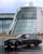A sleek black Mercedes-Benz car drifts on a wet track in front of the iconic Mercedes-Benz World building, featuring a curved glass façade and large logo.