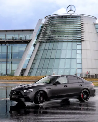 A sleek black Mercedes-Benz car drifts on a wet track in front of the iconic Mercedes-Benz World building, featuring a curved glass façade and large logo.