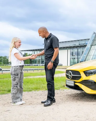 Young girl shaking hands with a Mercedes-Benz instructor outside Mercedes-Benz World, next to a bright yellow vehicle, symbolizing a positive learning experience.