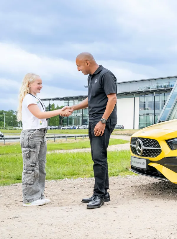 Young girl shaking hands with a Mercedes-Benz instructor outside Mercedes-Benz World, next to a bright yellow vehicle, symbolizing a positive learning experience.