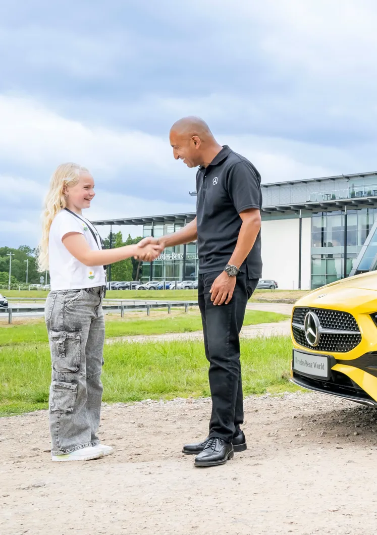 Young girl shaking hands with a Mercedes-Benz instructor outside Mercedes-Benz World, next to a bright yellow vehicle, symbolizing a positive learning experience.