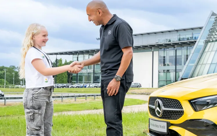 Young girl shaking hands with a Mercedes-Benz instructor outside Mercedes-Benz World, next to a bright yellow vehicle, symbolizing a positive learning experience.