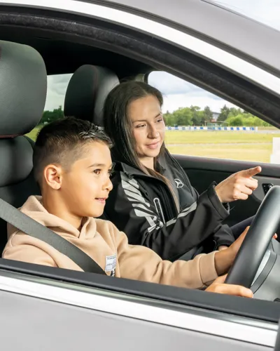 Young boy driving a car with a female instructor beside him, guiding and pointing ahead during a junior driving experience on a track.