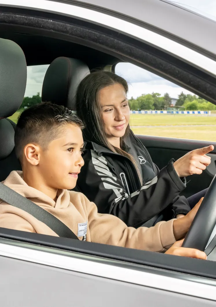 Young boy driving a car with a female instructor beside him, guiding and pointing ahead during a junior driving experience on a track.