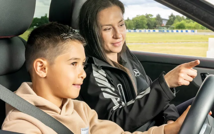 Young boy driving a car with a female instructor beside him, guiding and pointing ahead during a junior driving experience on a track.