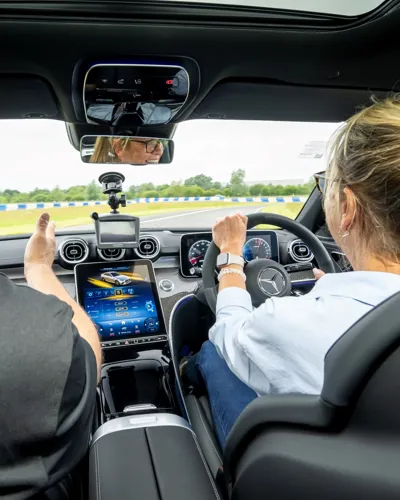 Interior view of a Mercedes-Benz during a driving experience, with a woman at the wheel, instructor beside her, and advanced dashboard and navigation systems visible.