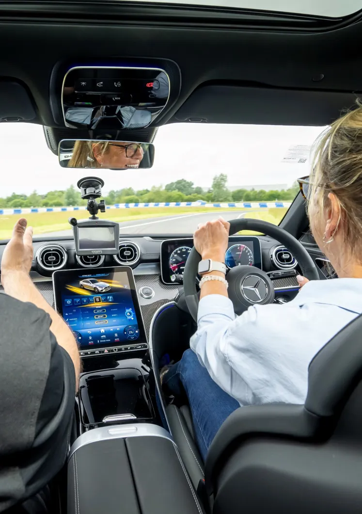 Interior view of a Mercedes-Benz during a driving experience, with a woman at the wheel, instructor beside her, and advanced dashboard and navigation systems visible.
