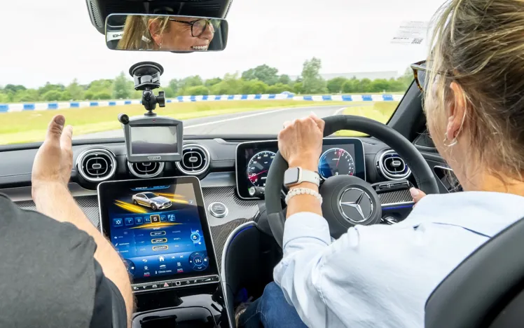 Interior view of a Mercedes-Benz during a driving experience, with a woman at the wheel, instructor beside her, and advanced dashboard and navigation systems visible.